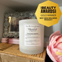 Candle labeled 'Flourish' with a decorative mirror and pink flower in the background, featuring a 'Gold Winner Best New Wellness Product' badge.